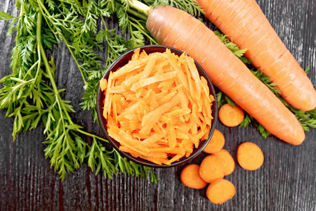 Grated carrots in a bowl, whole with haulm, sliced, brown napkin on the background of a wooden board from aboveの写真素材