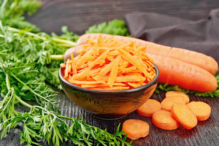 Grated carrots in a bowl, whole with haulm, sliced, brown napkin on the background of a wooden boardの写真素材