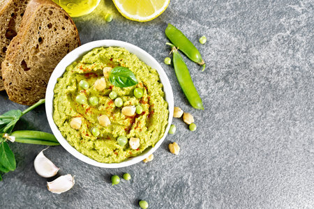 Hummus from green peas and chickpeas in a bowl, pea pods, lemon, garlic, vegetable oil in a carafe and slices of bread on granite countertop background from aboveの写真素材