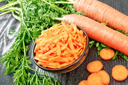 Grated carrots in a bowl, whole with haulm, sliced, brown napkin on the background of a dark wooden boardの写真素材
