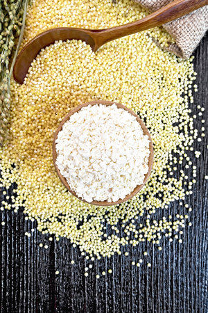 Millet flakes in a bowl, groats on the table, in a spoon and a bag of burlap, stalks with ears of millet on a background of wooden board from aboveの写真素材