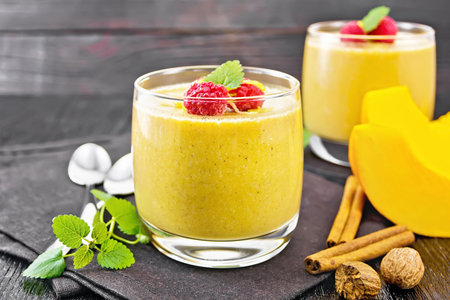 Yogurt, pumpkin and raspberry dessert with nutmeg, cinnamon, orange zest in a glass, vegetable, spoons and spices on a napkin on a dark wooden board backgroundの写真素材