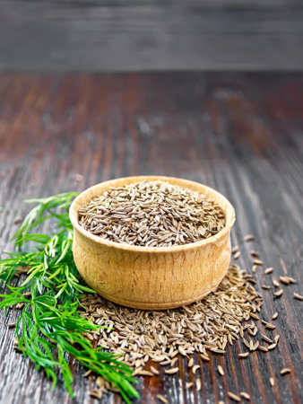 Cumin seeds in a bowl and on table, a green twig of plant on dark wooden board backgroundの写真素材