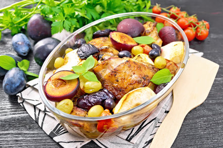 Baked chicken with tomatoes, apples, plums and grapes in a glass roaster on a towel, garlic, parsley and basil on wooden board backgroundの写真素材