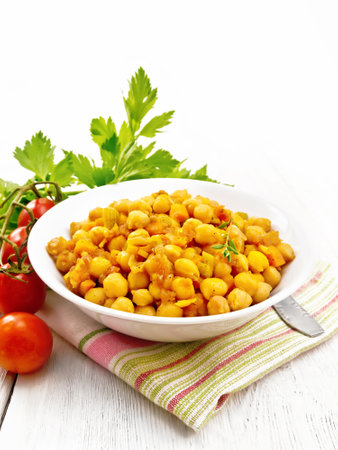 Stewed chickpeas with celery, tomatoes, onions, carrots and garlic in a plate on a towel against the background of light wooden boardの写真素材