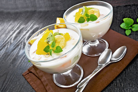 Dessert airy yogurt with orange, banana and kiwi in two glass bowls, fruits, spoons and mint on a brown towel on background of wooden boardの写真素材