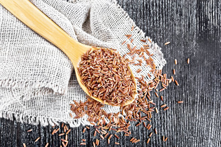 Red raw rice in a spoon on a burlap napkin on background of wooden board from aboveの写真素材