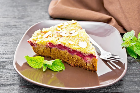 A piece of sweet cake with raspberries, butter crumble and almond flakes in a plate, mint and napkin on dark wooden board backgroundの写真素材