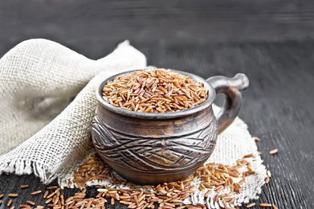 Red raw rice in a clay cup on a burlap napkin against dark wooden boardの写真素材