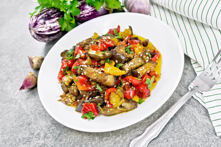 Vegetable stew with eggplant, sweet pepper, onion, fried with herbs, garlic and spices in a plate, towel, parsley on background of a gray granite tableの写真素材