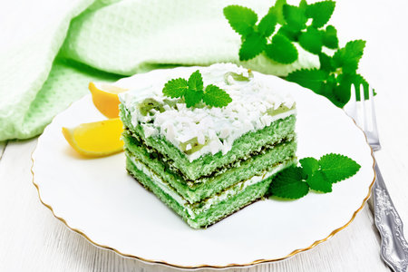 Piece of cake with mint and orange, white cream, confit, green sponge cake, sprinkled with white chocolate and green jelly in a plate, towel on wooden board backgroundの写真素材