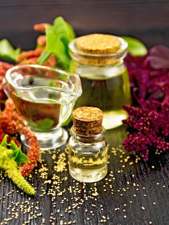 Amaranth oil in a glass bottle, gravy boat and jar, brown, green and purple flowers and leaves of the plant on dark wooden board backgroundの写真素材
