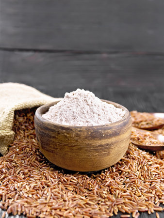 Red rice flour in bowl, cereal in bag, two spoons and on table on the background of wooden boardの写真素材