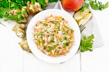 Salad of Jerusalem artichoke, apple, carrot and stalked celery with cream in plate, vegetable tubers, napkin on background of wooden board from aboveの写真素材