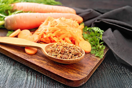 Dried carrots in a spoon, whole with haulm and grated, brown napkin on the background of a dark wooden boardの写真素材