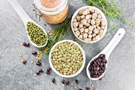 Green lentils and chickpeas in bowls, ruby haricot and mung beans in spoons, red lentils in a glass jar, thyme sprigs on a stone tabletop background from aboveの写真素材