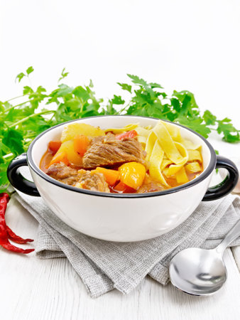 Central Asian national dish Lagman of meat, noodles and vegetables in bowl on kitchen towel, garlic, parsley on light wooden boardの写真素材