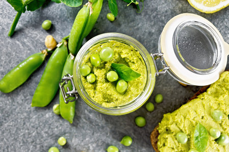 Hummus from green peas and chickpeas in glass jar, pea pods, lemon on granite countertop background from aboveの写真素材