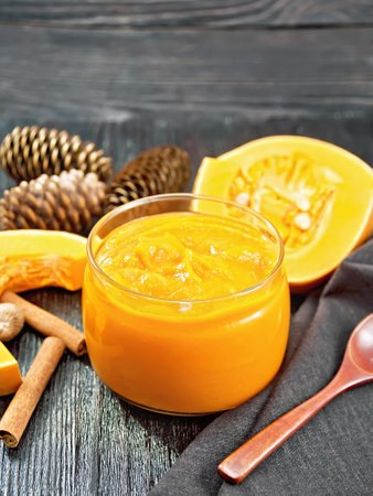 Pumpkin puree in a glass jar, pieces of raw vegetable. cinnamon, nutmeg, fir cones, spoon and napkin on dark wooden board backgroundの写真素材