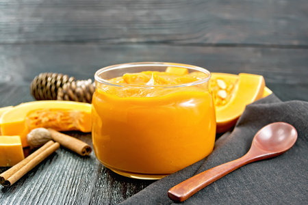 Pumpkin puree in a glass jar, pieces of raw vegetable. cinnamon, nutmeg, fir cones, spoon and brown napkin on a wooden board backgroundの写真素材