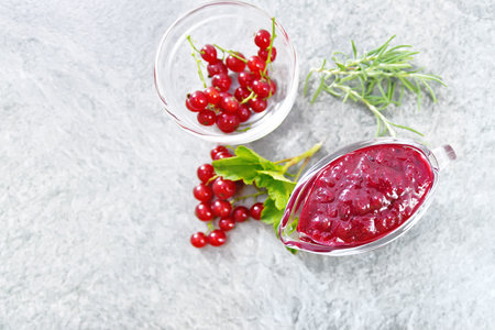 Redcurrant sauce in a glass sauceboat, berries in a bowl and on the table, a sprig of rosemary on a stone table background from aboveの写真素材