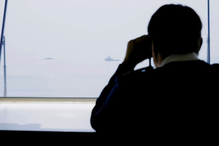 A man from his cabin looks through binoculars at ships in the distance.
