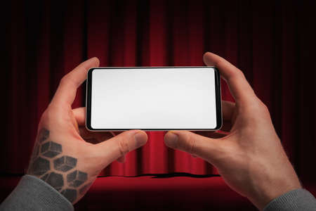Close up of Man Tattooed Hands Holding Modern Mobile Phone With White Blank Screen on Red Curtains Theatre Background. Concept of Online Cinema. Copy Space. Empty Space. 3d Rendering.の写真素材