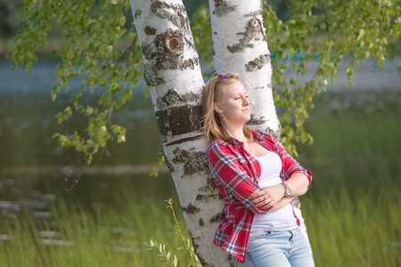 Portrait of a beautiful young woman resting near the lake leaning against a birch treeの写真素材