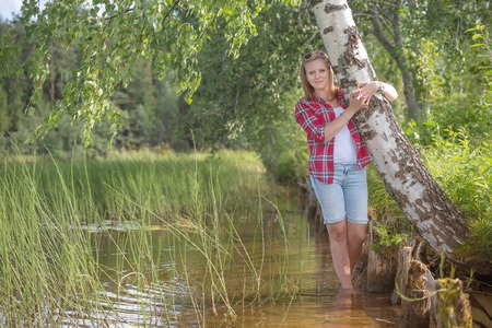 Portrait of a beautiful young woman standing in the water the lake leaning against a birch treeの写真素材