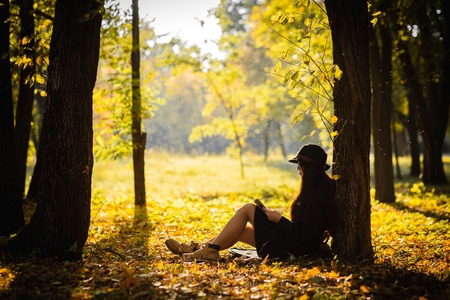 Girl in the hat in autumn park sits under a maple treeの写真素材
