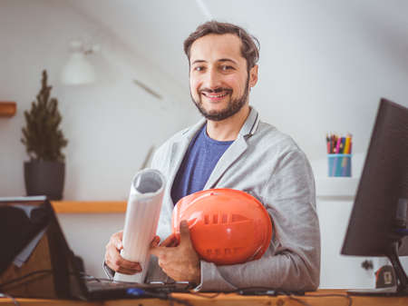 An attractive man with a beard smiles and poses for a photo with blueprints and a helmet sitting at a work table with computers. Color gradedの写真素材