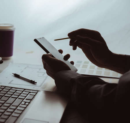 Silhouette of the hands of a man who works with a mobile phone at a table with a laptop coffee and drawingsの写真素材