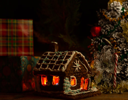 New Year's composition. A gingerbread house with lighting inside against the backdrop of gifts and a New Year's tree made of cones with tinsel decorations.の写真素材