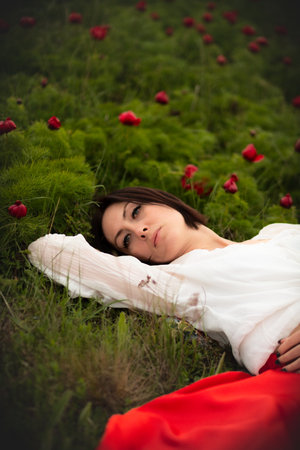 Artistic photo. Vertical composition. An adult young woman in a white blouse and red skirt lies in a green meadow with scarlet wild peonies. Summer bright landscape.の写真素材
