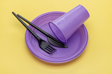 Disposable purple plates, cup, black knife and fork, yellow background. plastic recycling.