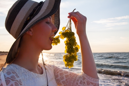 Beautiful young woman in hat atsea shore holding grapesの写真素材