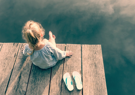 Adorable small girl sitting on wooden bridge and looking on river and sunの写真素材