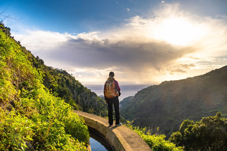 Tourist on Levada do Norte on the Portuguese island of Madeira. Levada irrigation canal. Hiking in Madeira. Narrow path next to the levada. Green mountains and ocean in background.の写真素材