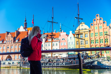 Tourist in red jacket with backpack walks along Gdansk waterfront. Behind her are historical buildings with vibrant facades and a traditional sailing ship at the harbor.の写真素材