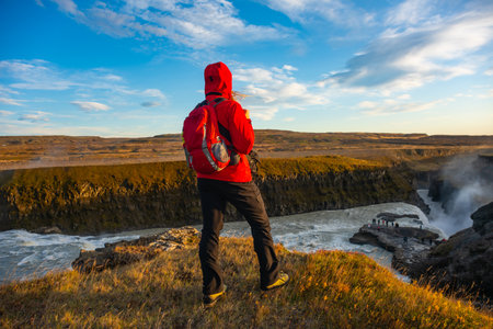 Adventurer stands triumphantly near a stunning Icelandic waterfall Gullfoss. Surrounded by rugged terrain, the person embraces the breathtaking natural beauty and vibrant rushing watersの写真素材