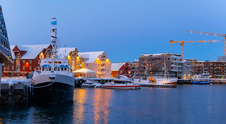 Evening scene at Tromso harbor featuring docked boats under a snowy landscape. Warm lights from red...