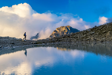 A person stands on rocky terrain, gazing at a stunning turquoise mountain lake surrounded. Dressed in hiking gear and holding trekking poles. Wildsee Pizol, Switzerland.の写真素材