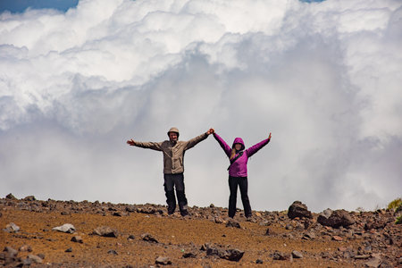 A couple stands together on rocky terrain, holding hands and smiling. The backdrop showcases fluffy clouds and a clear blue sky, creating a scenic outdoor moment.の写真素材