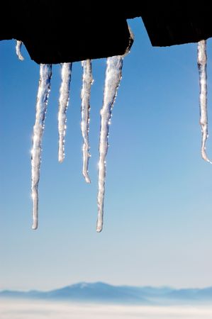 The icicles which are hanging down from a wooden roof, against the blue sky and mountains