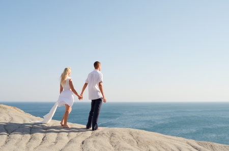Young couple dressed in white, holding hands and looking at the seaの写真素材