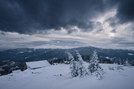 Bad weather in the mountains. Winter landscape. Cloudy evening with storm clouds. Wooden houses of shepherds under the snow. Carpathians, Ukraine, Europeの写真素材