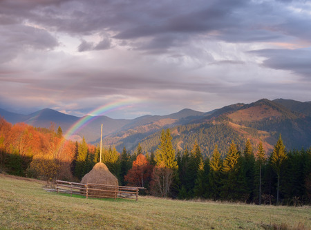 Autumn landscape in a mountain village. Haystack on the meadow. Rainbow over the hills. Collage of two frames. Art processing photosの写真素材