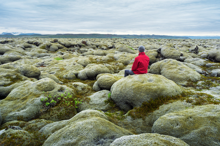 Traveler in a red jacket sits on a moss. Lava field on the south coast of Iceland, Europe. Tourist attraction. Amazing in nature. Cloudy summer dayの写真素材