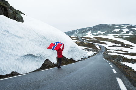Bjorgavegen - Snowy road in Norway. Girl with the Norwegian flag on a mountain road near a wall of snow. Severe northern landscapeの写真素材
