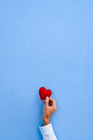 Red heart in a man's hand on blue wall background. Valentine's Day card with copy space for greeting text. Central symmetric compositionの写真素材